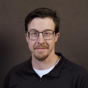 Carl Busch headshot in a black shirt with a grey background